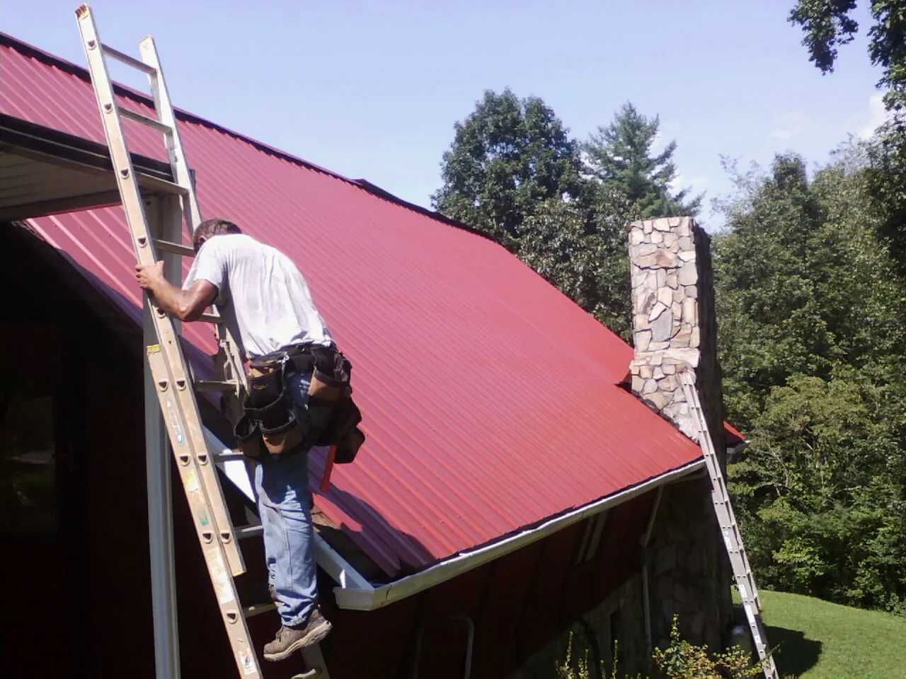 Roofer on a ladder beside a red metal roof and stone chimney — Soffit & Fascia Repair by Roofing Brothers in Weeping Water